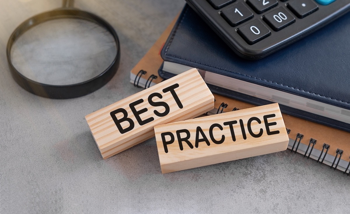 Wooden blocks labeled “BEST” and “PRACTICE” resting against a notebook and calculator with a magnifying glass beside them.