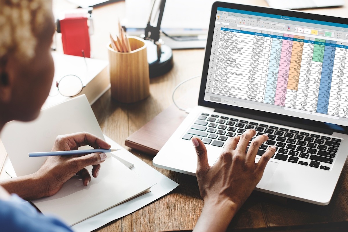 A person viewing a spreadsheet on a laptop while taking notes at a desk.