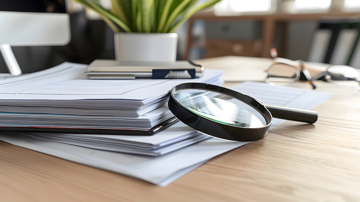 A stack of contracts and papers on a desk with a large magnifying glass resting on top.