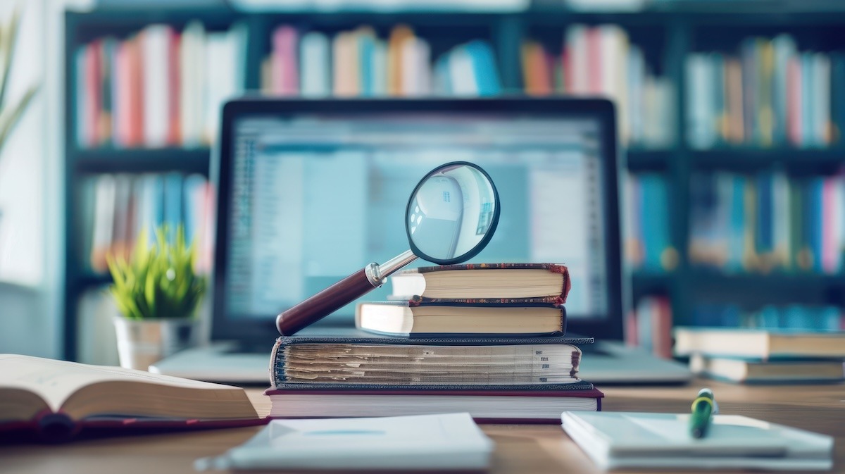 A stack of books with a magnifying glass on top, with papers, an open book, and a pen in the foreground and a laptop and library shelf blurred in the background.