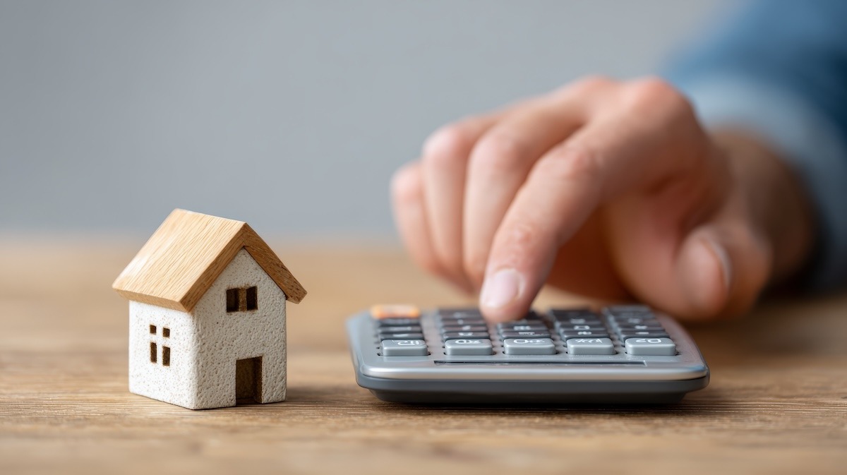 A close-up of a hand using a calculator with a small toy house beside it.