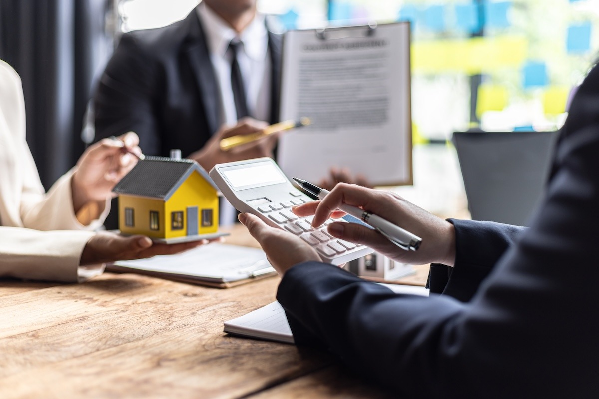 People seated at a desk with a calculator, documents on a clipboard, and a model house held in one hand.