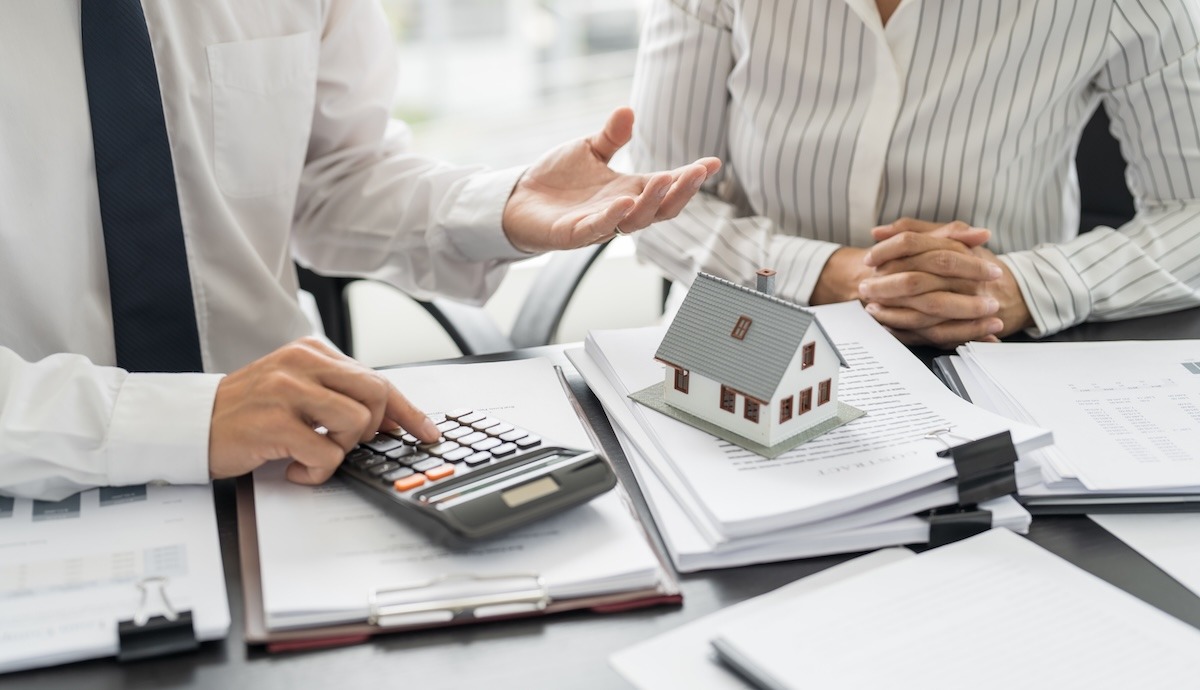 Two people reviewing contracts at a desk with one person using a calculator and a small model house on top of the papers.