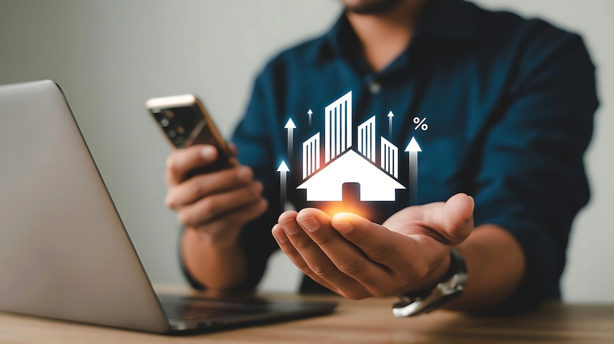 A person sitting at a desk with a phone in one hand and a graphic of a house with upward arrows and a percent symbol in the other hand.