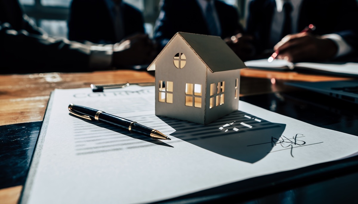 A small toy house sitting on a clipboard with a contract and a pen beside it.