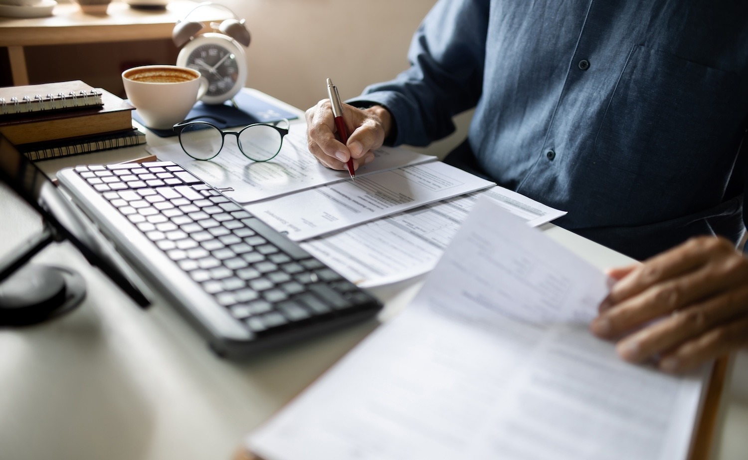 Person writing notes at a desk with a computer keyboard, glasses and coffee