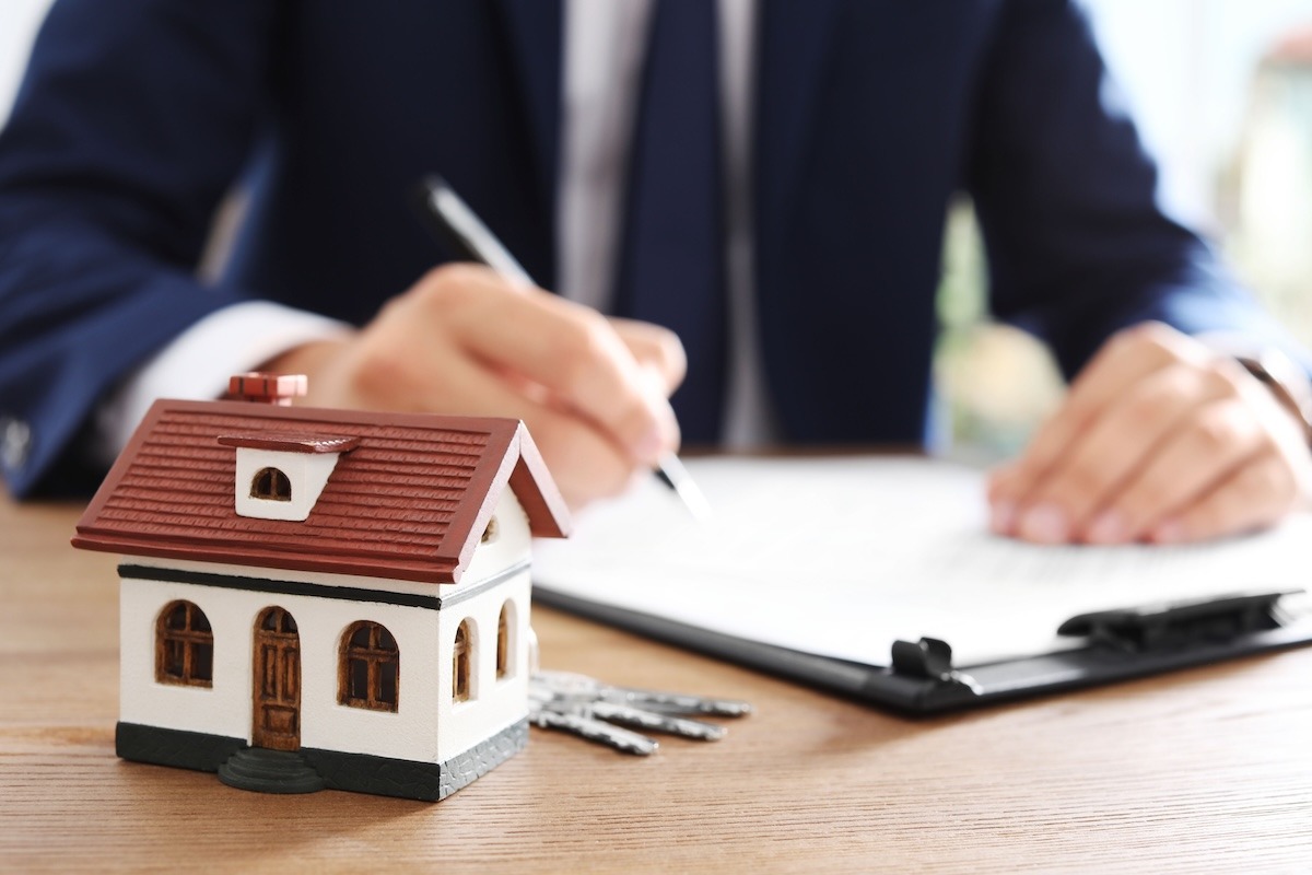A model house with keys beside a document in the foreground, with a blurred person in the background.