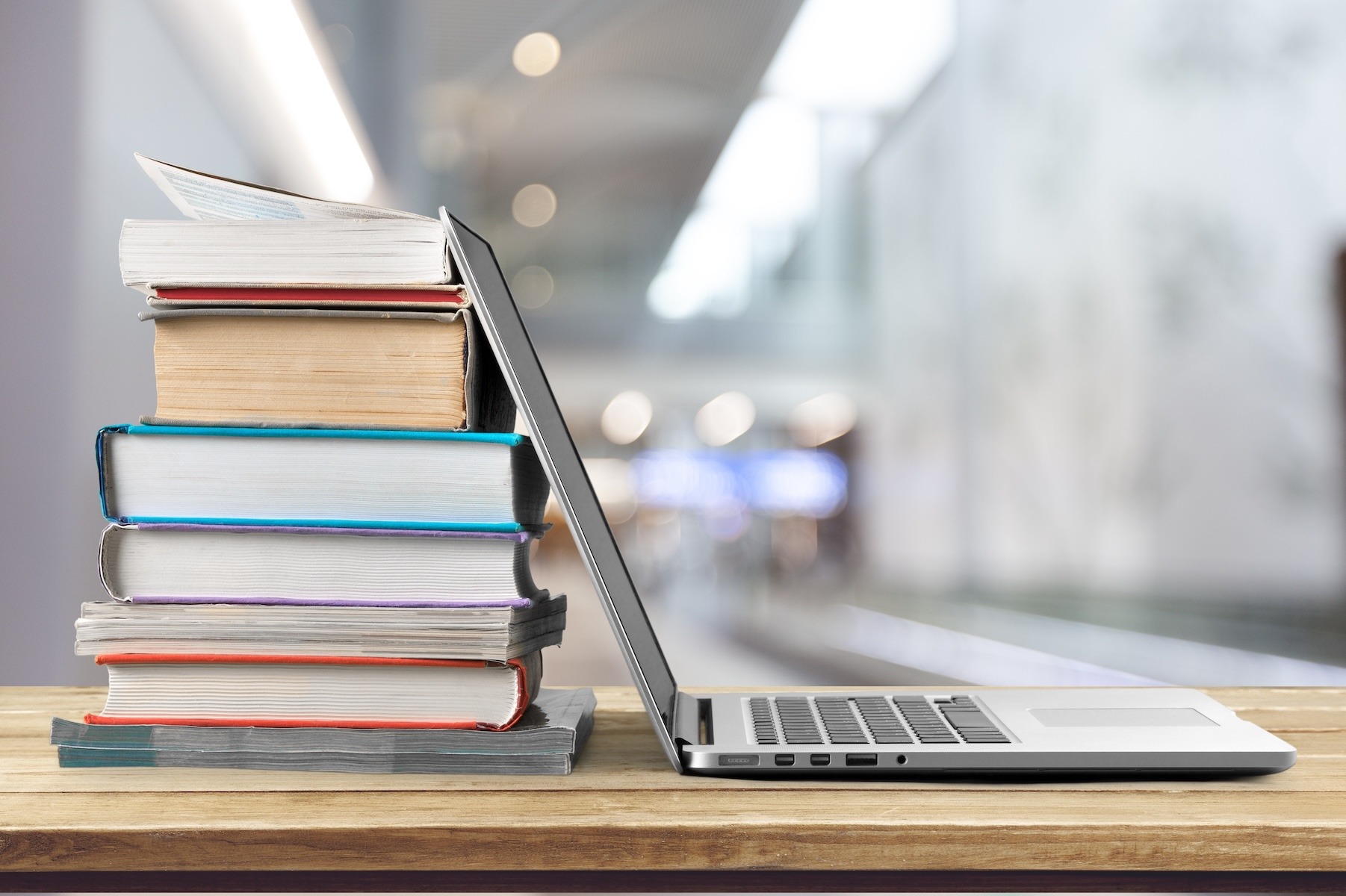 A stack of books with a laptop resting against them on a table, with shelves in the background.