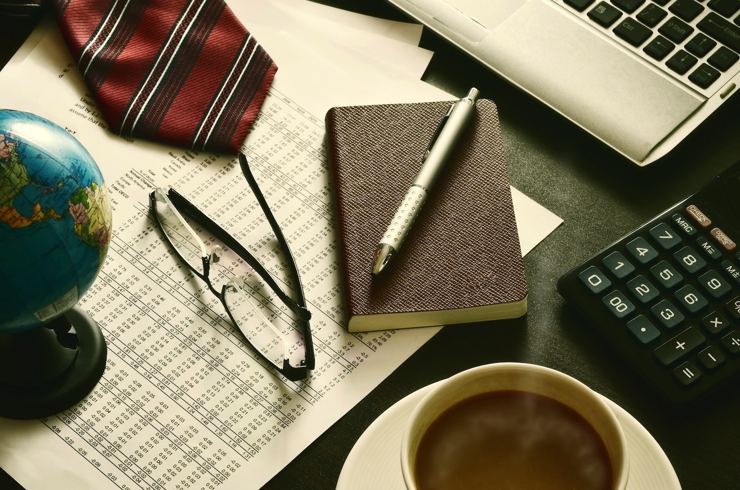 Desk with notebook, calculator, financial documents, glasses, laptop and coffee.