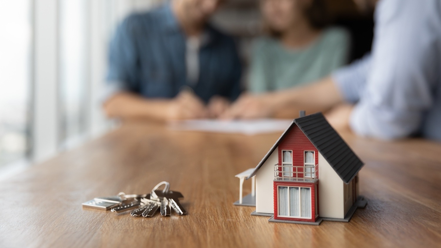 Model house and keys with people reviewing property documents in the background.
