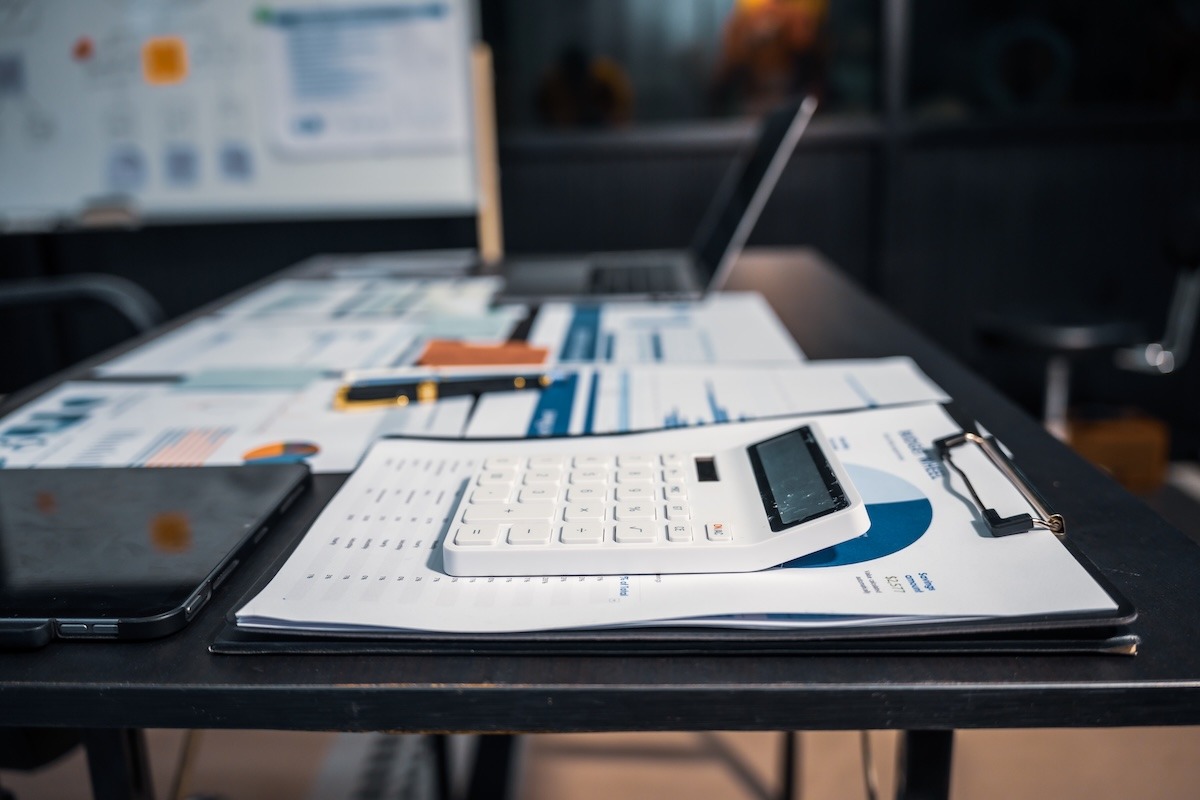 An organized office desk with a clipboard and calculator on the right, an iPad below it, papers on the left, and a laptop at the far end.